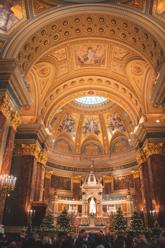 Interior of St. Stephen’s Basilica in Budapest beautifully lit during Christmas, showcasing its ornate golden dome, religious frescoes, and festive decorations with visitors admiring the altar.