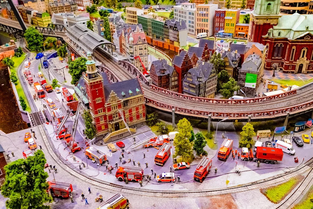 Miniature Wunderland model city with emergency fire trucks and ambulances responding to a scene near a train station and historic buildings, detailed scale model with urban architecture and elevated railway.