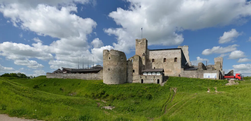 A medieval stone castle with rounded towers and a central tall structure, reminiscent of Rakvere Castle, sits atop a grassy hill under a blue sky with scattered clouds. A red truck is parked beside the castle.