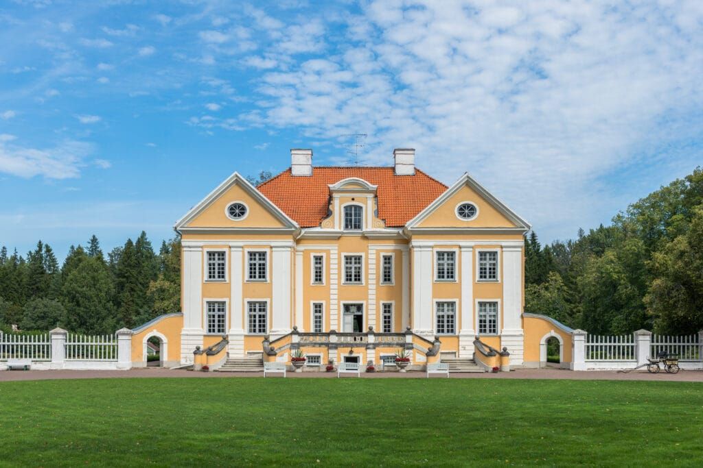 Front view of the elegant Baroque-style Palmse Manor in Lahemaa National Park, Estonia, with a bright yellow façade, red-tiled roof, and manicured green lawn under a blue sky.