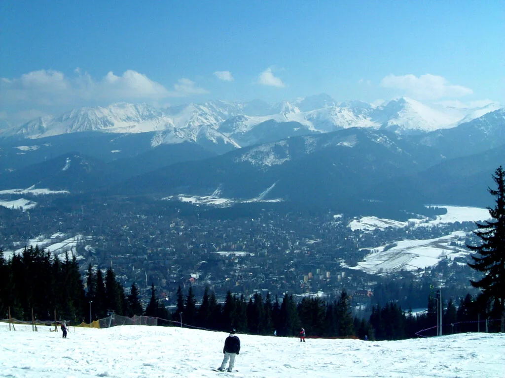 A picturesque winter scene shows Zakopane Poland from a Gubałówka Mountain ski slope where people ski through snow-covered terrain while enjoying the Tatra Mountains landscape.