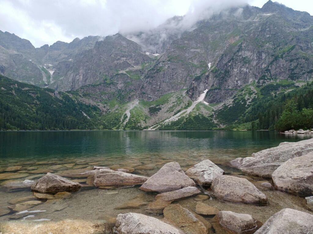 Scenic view of Morskie Oko Lake in the Tatra Mountains, Poland, with clear water, rocky shoreline, and misty mountain peaks in the background.