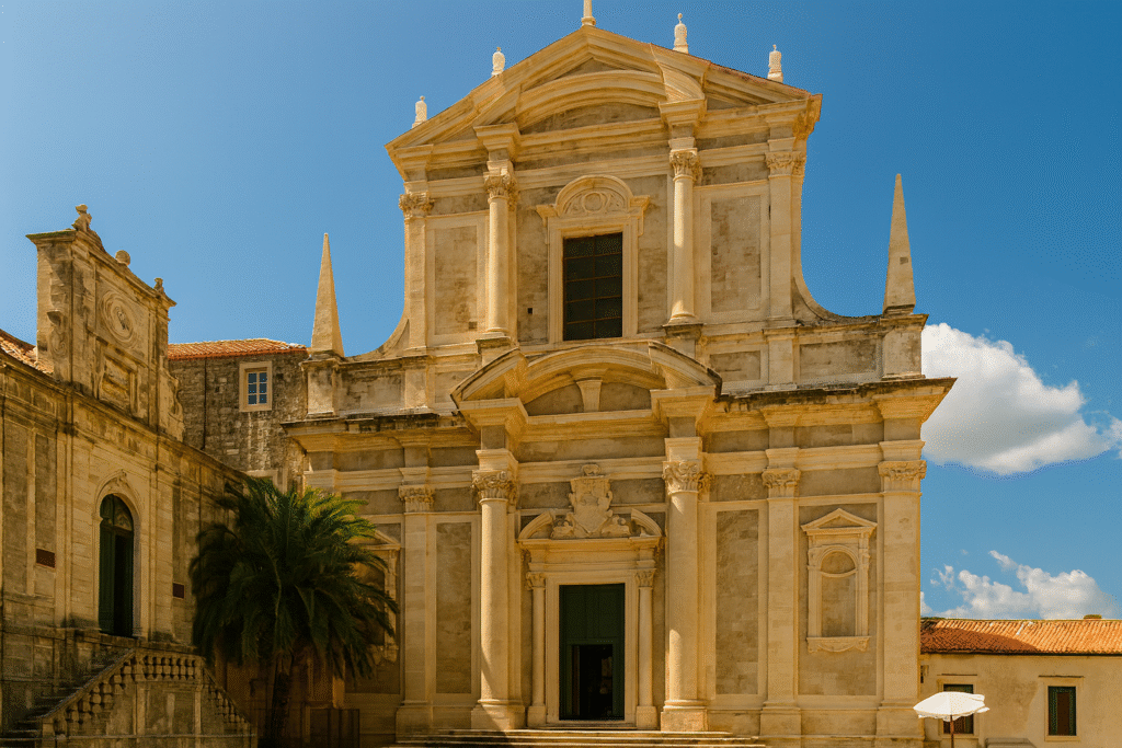 Baroque-style stone church facade with columns, statues, and detailed architecture against a bright blue sky with clouds