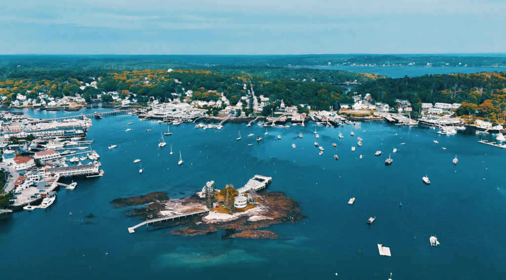 Aerial view of Boothbay Harbor, a picturesque coastal stop along the Portland Maine to Bar Harbor route, showing sailboats, docks, waterfront homes, and lush green scenery surrounding the harbor.