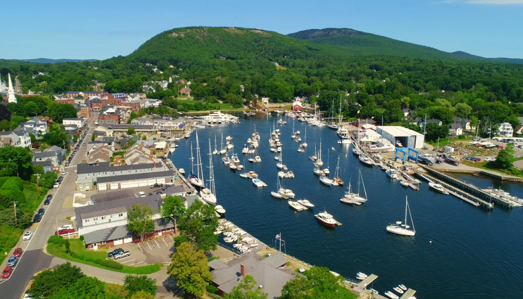 A scenic aerial view of Camden Harbor, a charming coastal stop along the Portland Maine to Bar Harbor route, featuring sailboats, waterfront buildings, and lush green hills in the background.