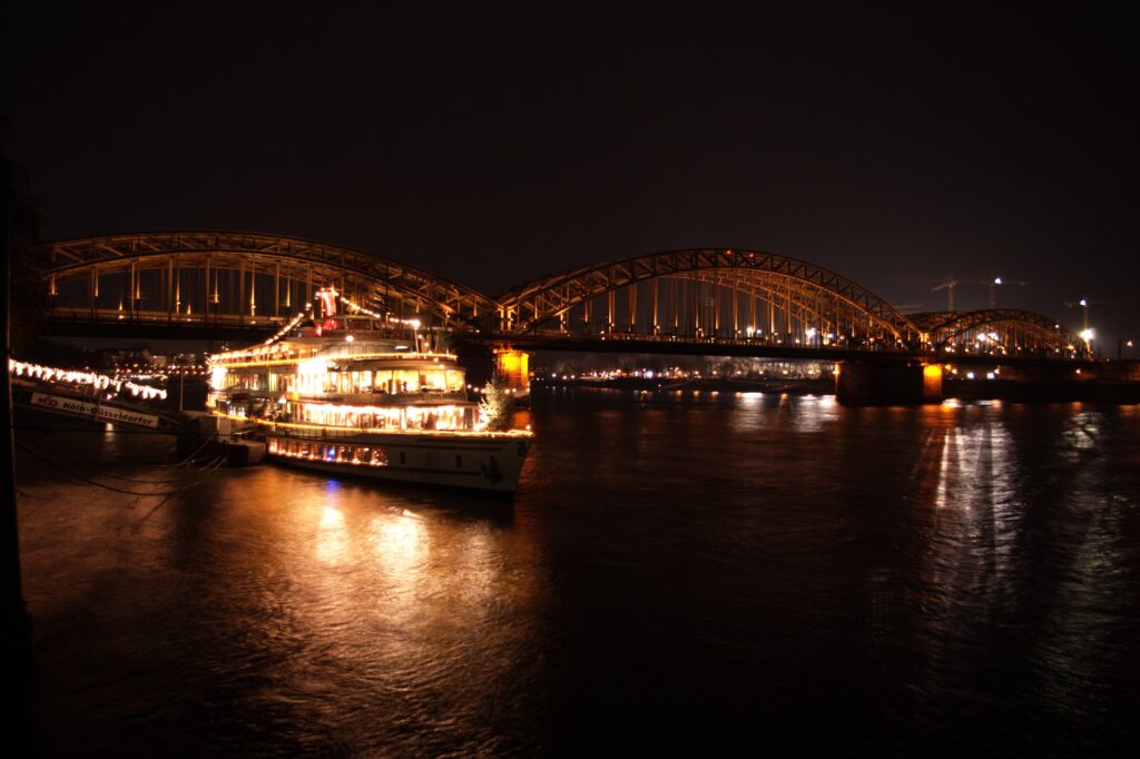 Night rhine river cruise on the Rhine River in Cologne, Germany, with city skyline and illuminated waterfront buildings at sunset.