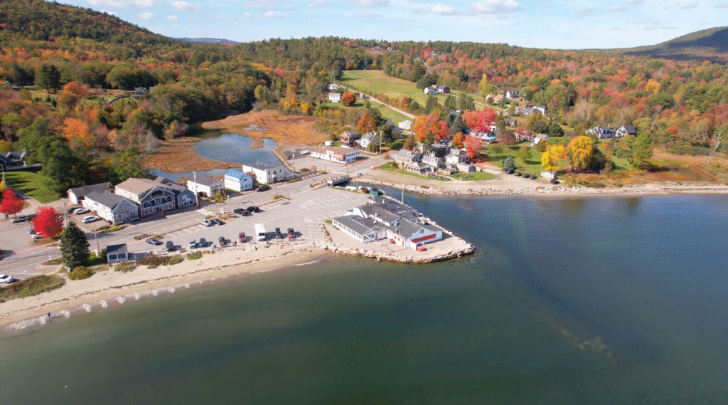 Aerial view of Lincolnville Beach along the scenic coastal route from Portland, Maine to Bar Harbor, showcasing sandy shoreline, waterfront buildings, parked cars, and vibrant fall foliage spread across rolling hills.