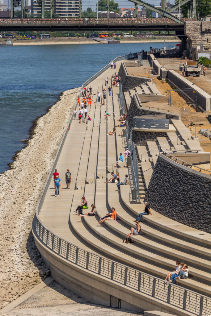 People walking and sitting along the Rhine River promenade in Cologne, Germany, during a period of low water levels exposing the rocky Rhine River bank.