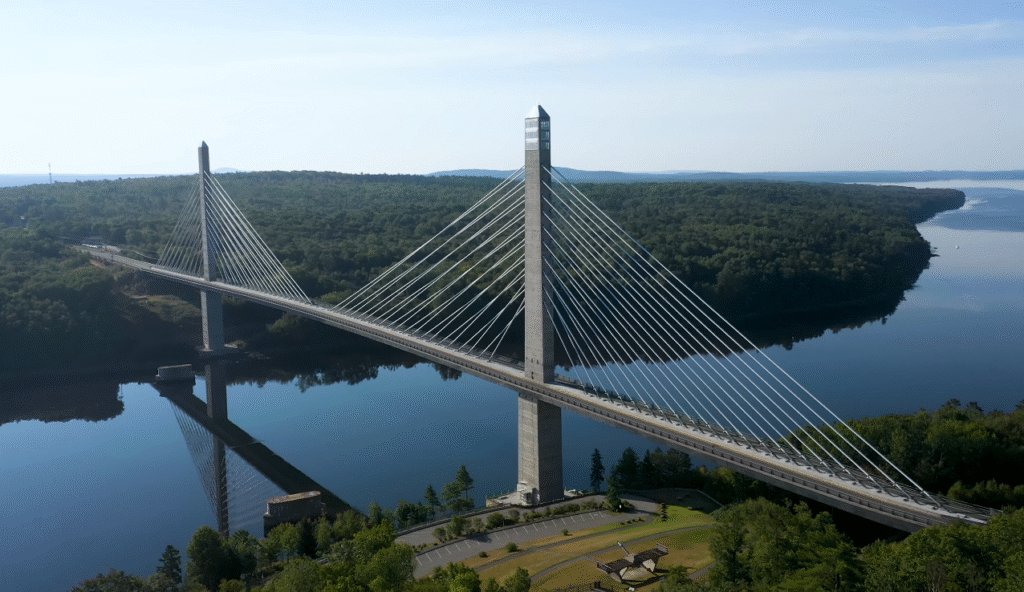 Aerial view of the Penobscot Narrows Bridge, a scenic landmark along the Portland Maine to Bar Harbor drive, spanning over calm river waters surrounded by lush green forests.