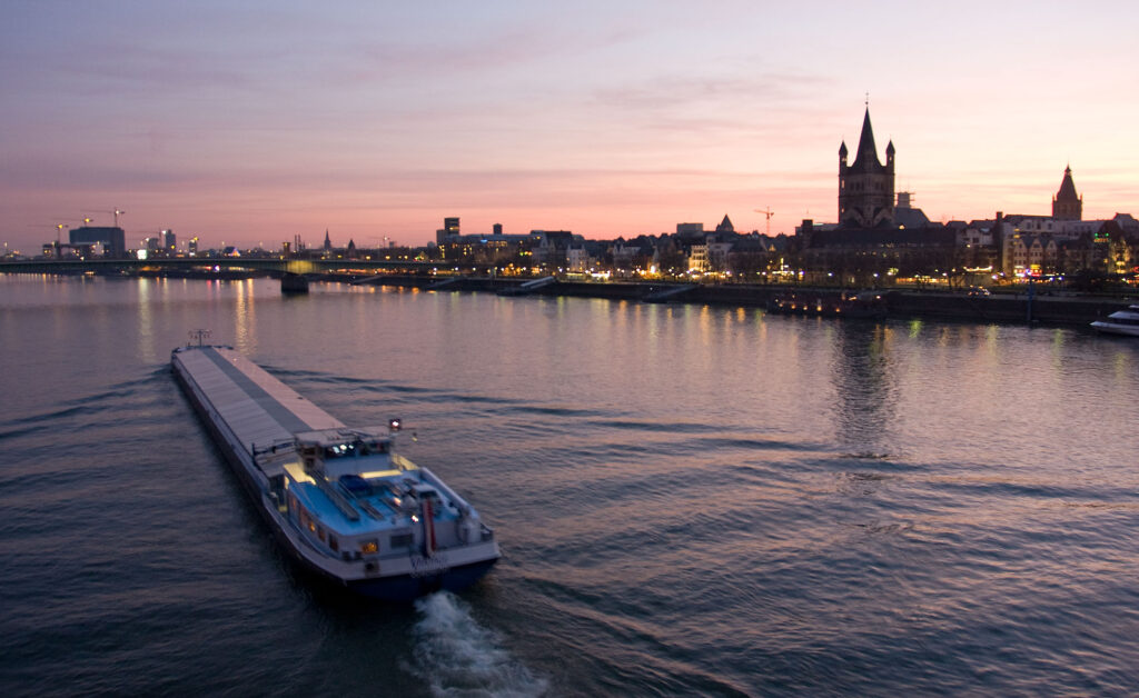 Evening rhine river cruise on the Rhine River in Cologne, Germany, with city skyline and illuminated waterfront buildings at sunset.