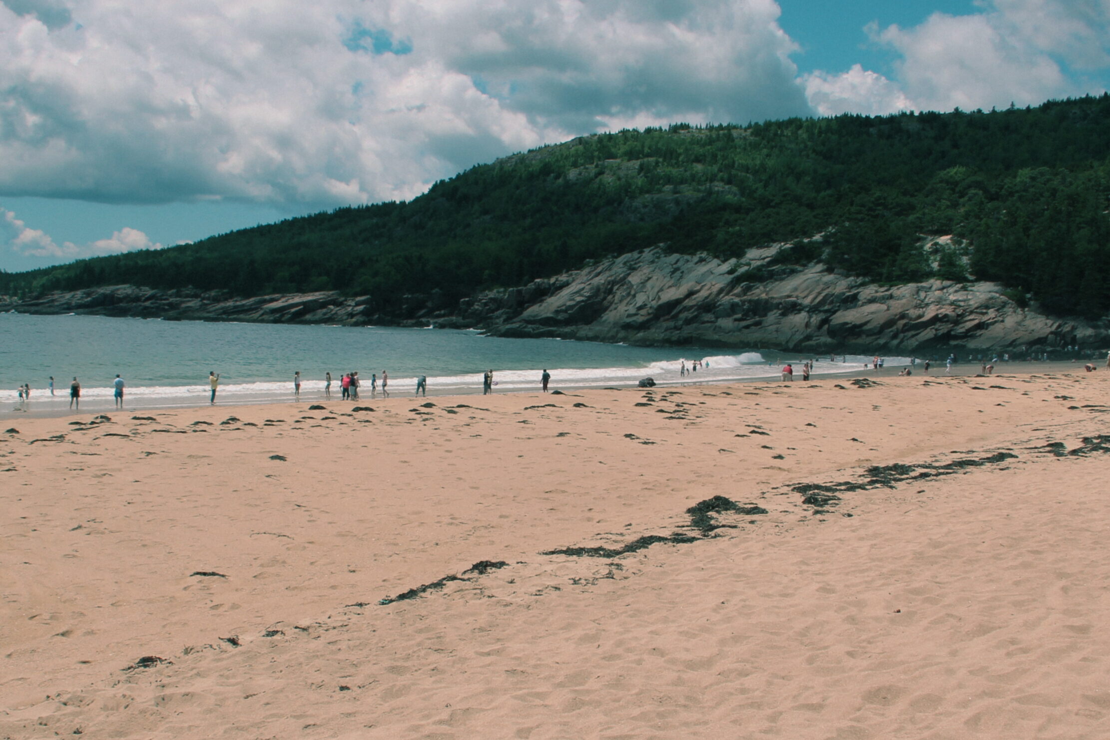 Wide sandy shoreline at Sand Beach in Acadia National Park with visitors enjoying the ocean and scenic mountain backdrop on a partly cloudy summer day – one of the top things to do in Acadia National Park.