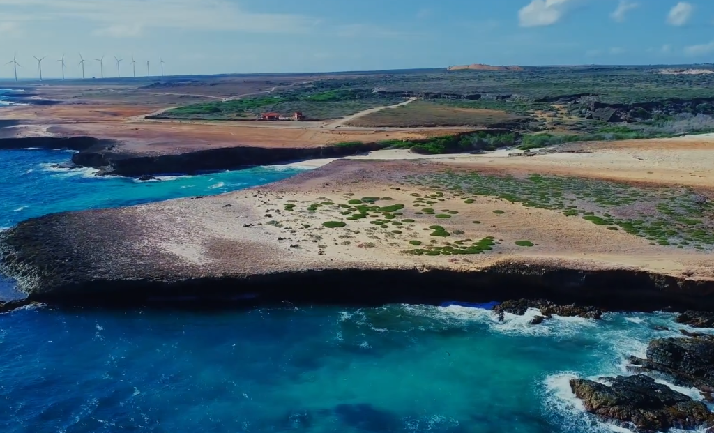 Aerial view of the rugged coastline at Arikok National Park showcasing the Natural Bridge area in Aruba, with turquoise waves crashing against rocky cliffs and desert landscape in the background