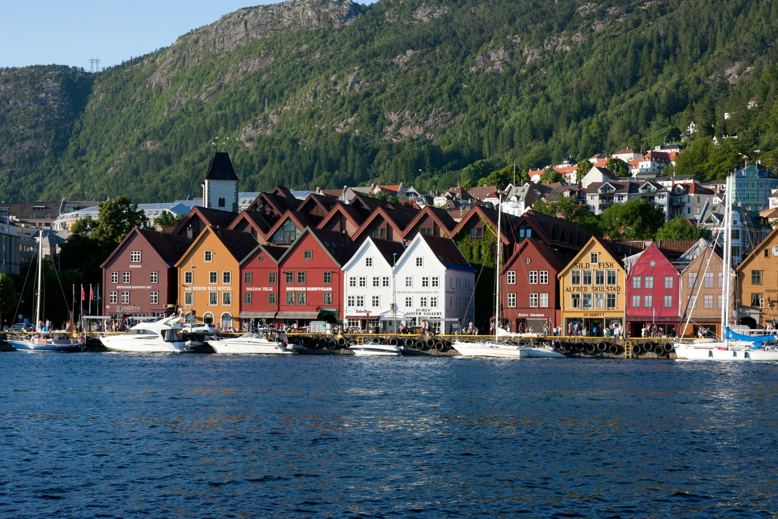 Scenic view of Bergen’s Bryggen waterfront with traditional wooden buildings, boats, and forested mountains, highlighting the charm of one of the largest cities in Norway