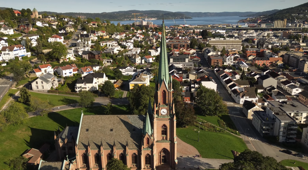 Drone view of a historic church and surrounding residential neighborhood in Drammen, Norway, with the river and hills in the background, showcasing the urban landscape connected to the largest cities in Norway such as Oslo, Bergen, Trondheim, and Stavanger