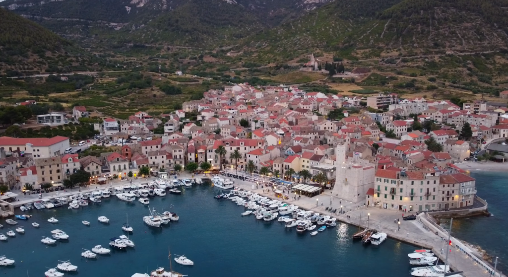 Aerial view of Komiža on Vis Island, Croatia, featuring the harbor filled with boats, historic stone houses with red roofs, and surrounding green hills at dusk