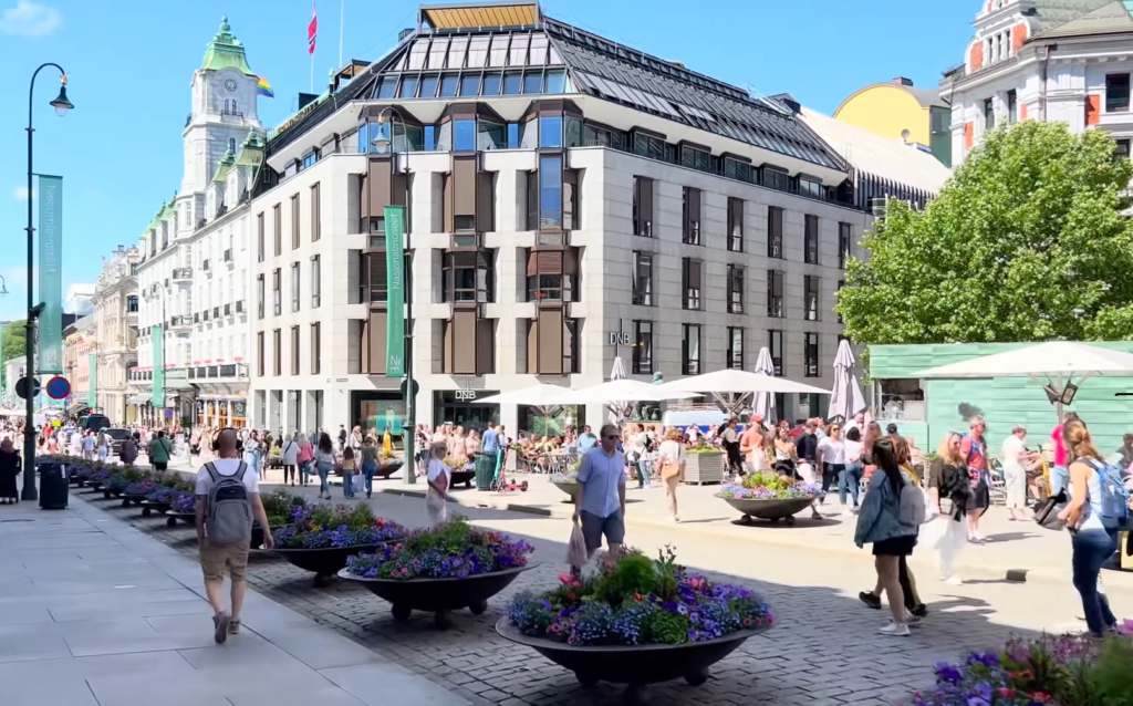 largest cities in Norway Busy street along Karl Johans gate, with people walking and sitting near large flower planters; modern buildings and outdoor café seating under umbrellas on a sunny day.