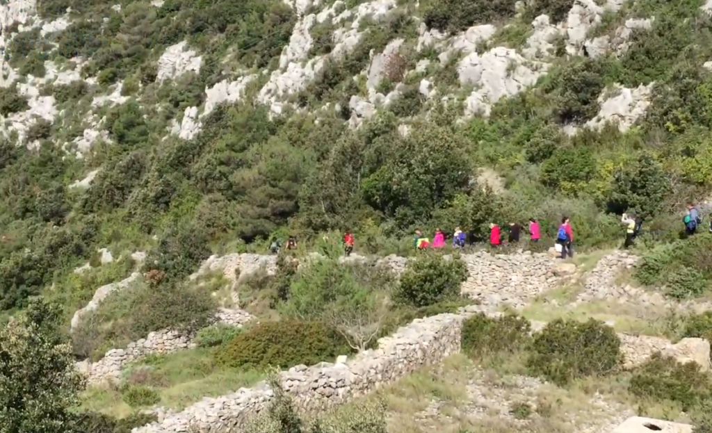 A group of hikers with colorful jackets walk along a narrow, winding trail through a Mount Hum Hike in vis island, green hillside. The scene conveys adventure and exploration.