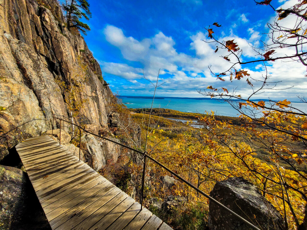 Dramatic cliffside boardwalk on the Precipice Trail in Acadia National Park, featuring iron rungs, steep granite cliffs, autumn foliage, and panoramic ocean views—one of the most thrilling Acadia hikes