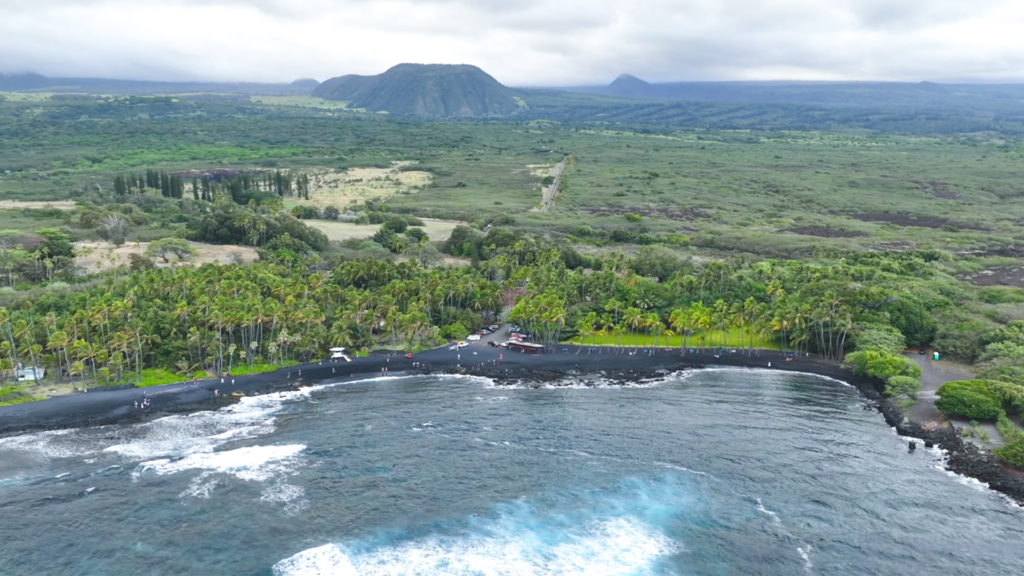 Aerial view of Punaluʻu Black Sand Beach in Hawaii showing volcanic black sand, palm trees, and Pacific Ocean coastline