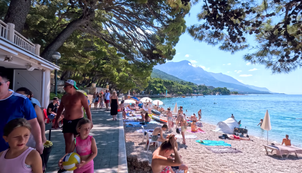 Crowded summer day at Punta Rata Beach in Brela, Croatia, with families relaxing along the pebble shoreline and clear blue water