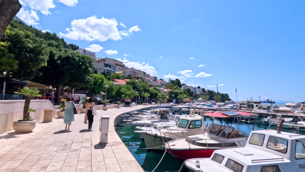 Coastal walking path near Punta Rata Beach in Brela, Croatia, with boats docked along the marina and people strolling by the waterfront