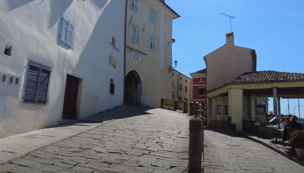 Steep stone pathway leading up to the historic hilltop entrance of Motovun, Croatia, with medieval buildings, outdoor cafés, and sunny blue skies