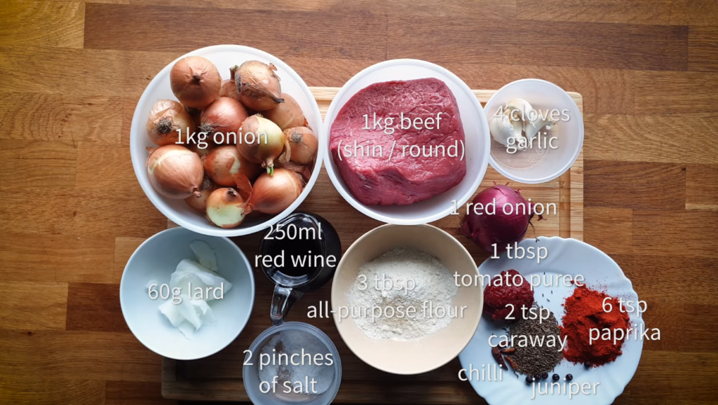 Flat lay of all ingredients - beef chunks, onions, paprika, caraway seeds, garlic, and spices arranged on a wooden surface