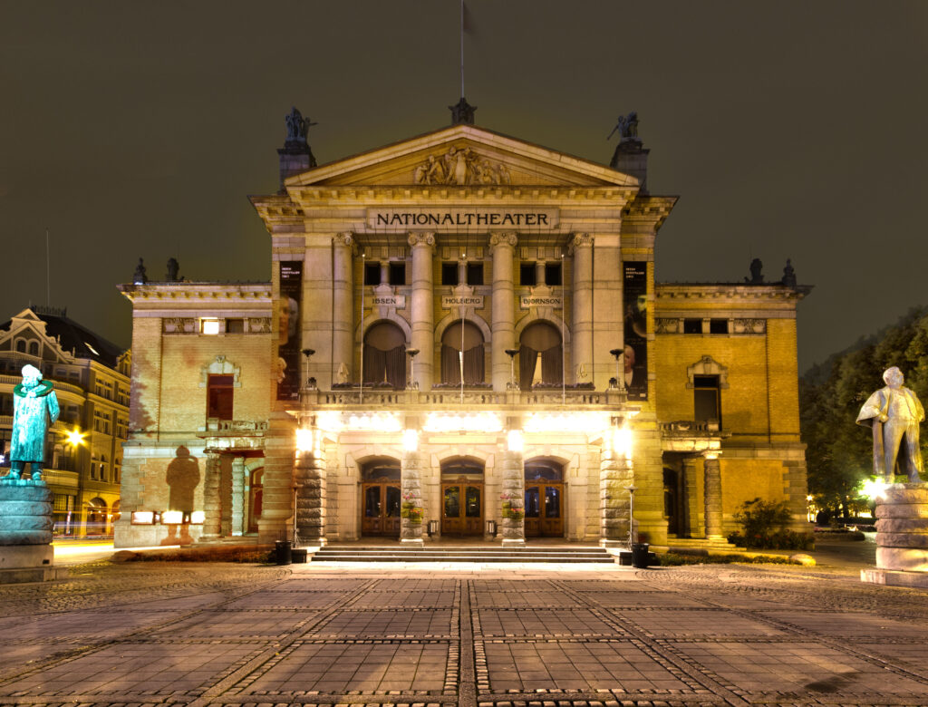 Illuminated at night, the grand facade of "Nationaltheater" near Karl Johans gate is flanked by statues, exuding a historic and elegant ambiance under a dim sky.