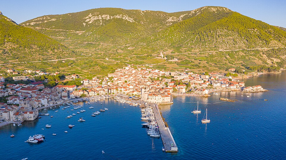 Aerial view of Vis Island, Croatia, showcasing the harbor, boats, and red-roofed town surrounded by lush green hills at sunset