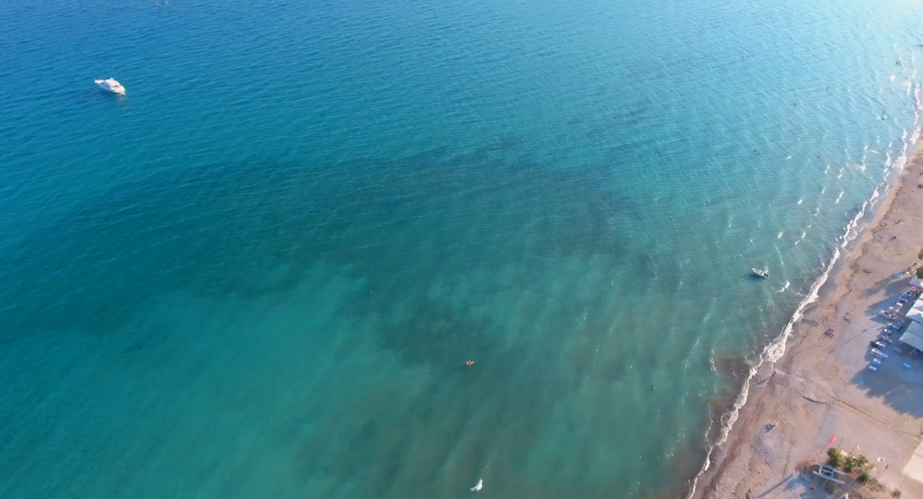 Aerial view of the calm turquoise sea at Varkiza Beach, with a small boat and sandy shoreline on the right, featuring some buildings and scattered beachgoers—one of the best beaches near Athens known for its sand and water quality.