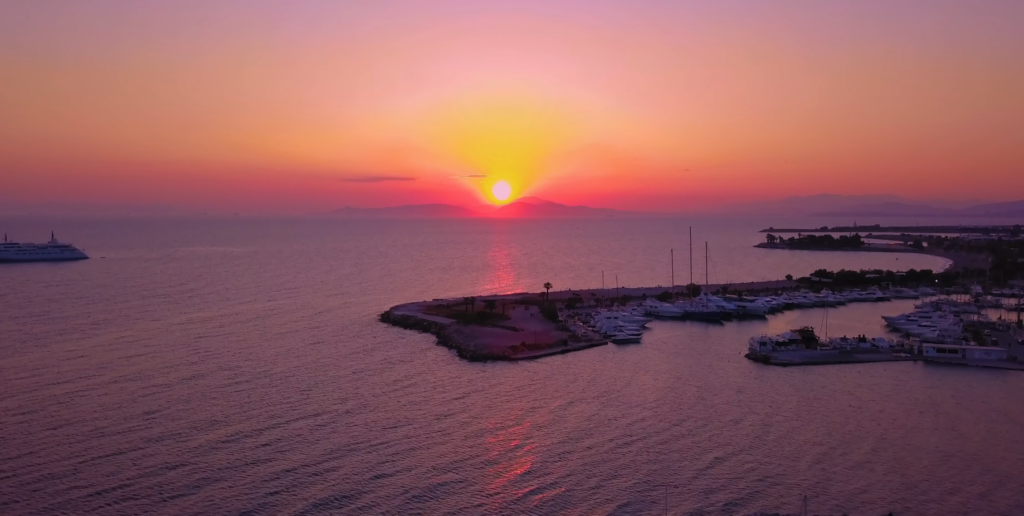 The sun sets over Varkiza Beach marina, casting a warm glow on a boat anchored in the calm waters